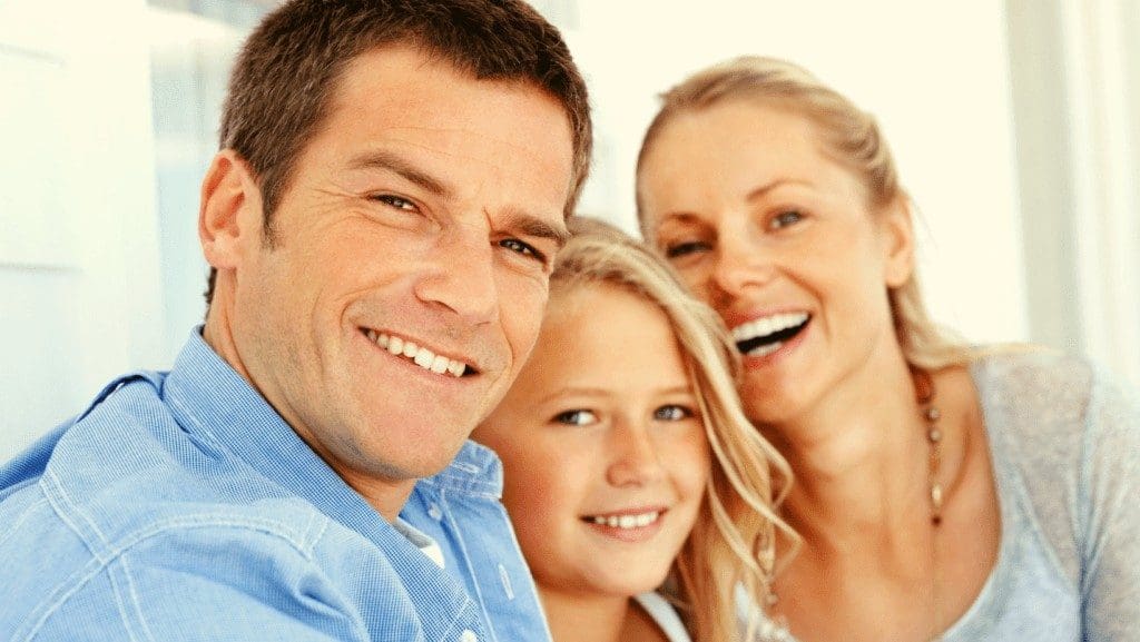 Man and woman smiling with dental bridges in Northampton and sitting on couch with their daughter