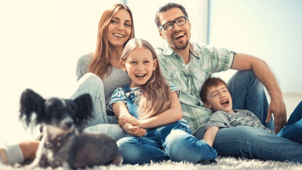 Family of four smiling and sitting on carpet with their dog