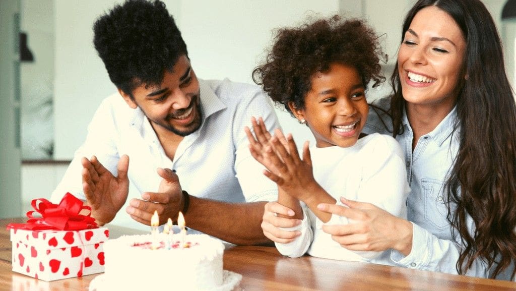 Laughing family of three sitting at table with small birthday cake and present