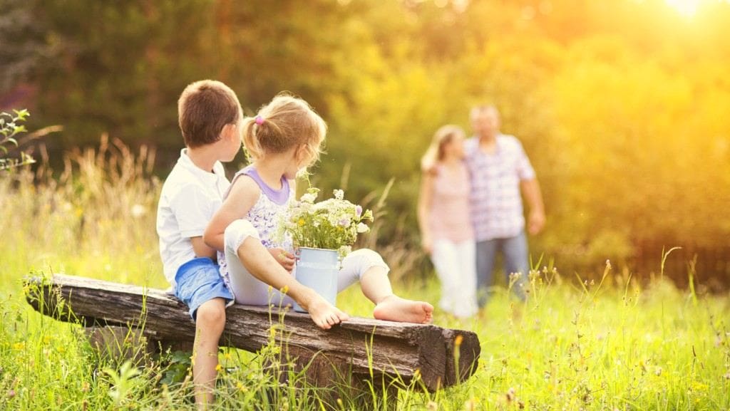 Two kids sitting on log outdoors with their parents walking in the background